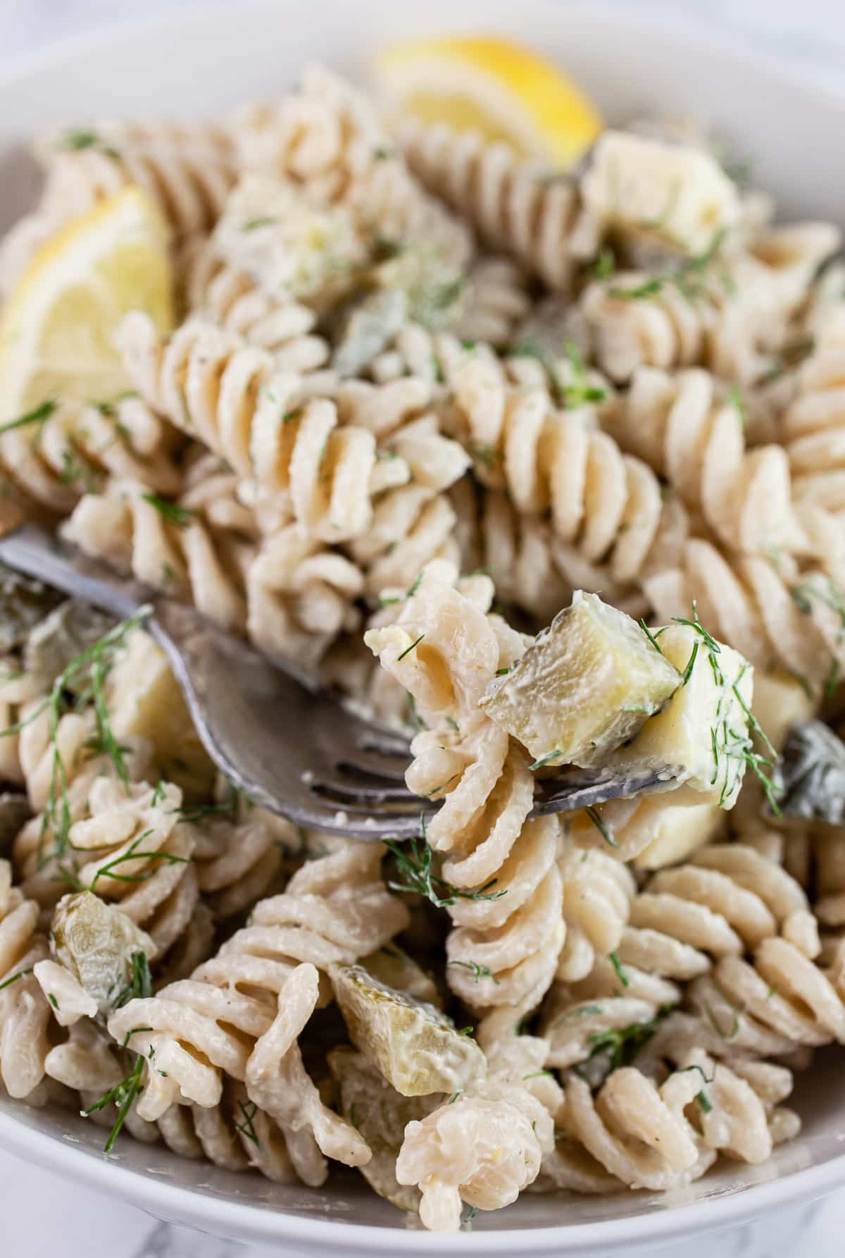 Dill pickle pasta salad in white bowl with fork and lemon wedge.