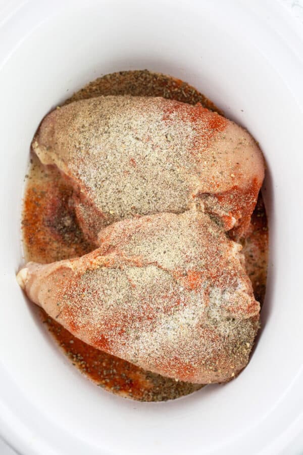 Uncooked split chicken breasts topped with spices in slow cooker.