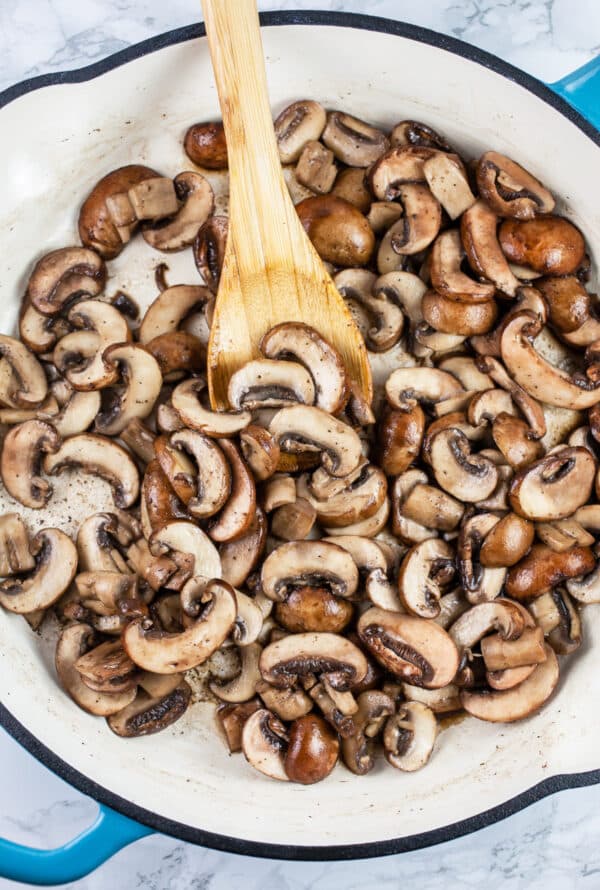 Sliced mushrooms sautéed in a skillet with wooden spoon.