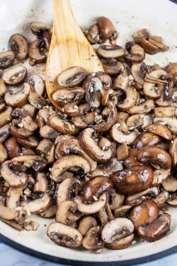 Sliced mushrooms and garlic sautéed in skillet with wooden spoon.