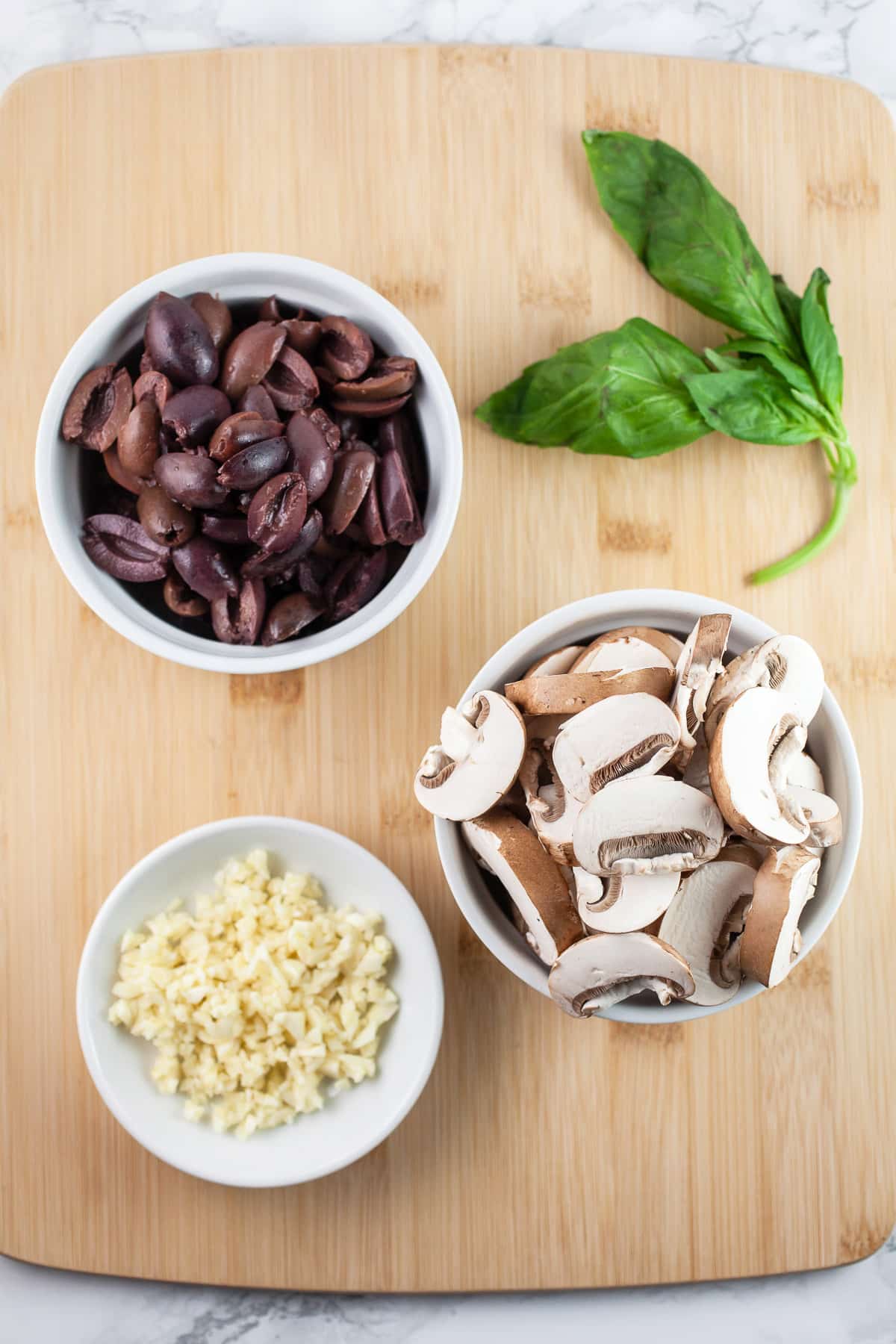 Minced garlic, diced Kalamata olives, sliced mushrooms, and fresh basil on wooden cutting board.