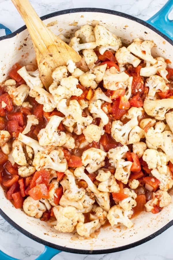 Cauliflower florets and diced tomatoes sautéed in skillet with wooden spoon.