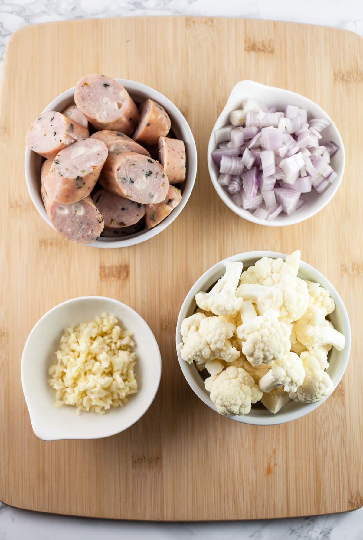Minced garlic, shallots, cauliflower florets, and sliced chicken sausages in white bowls on wooden cutting board.
