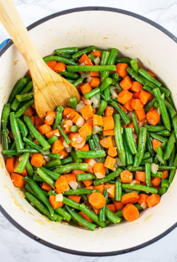Chopped carrots and green beans sautéed in Dutch oven with wooden spoon.