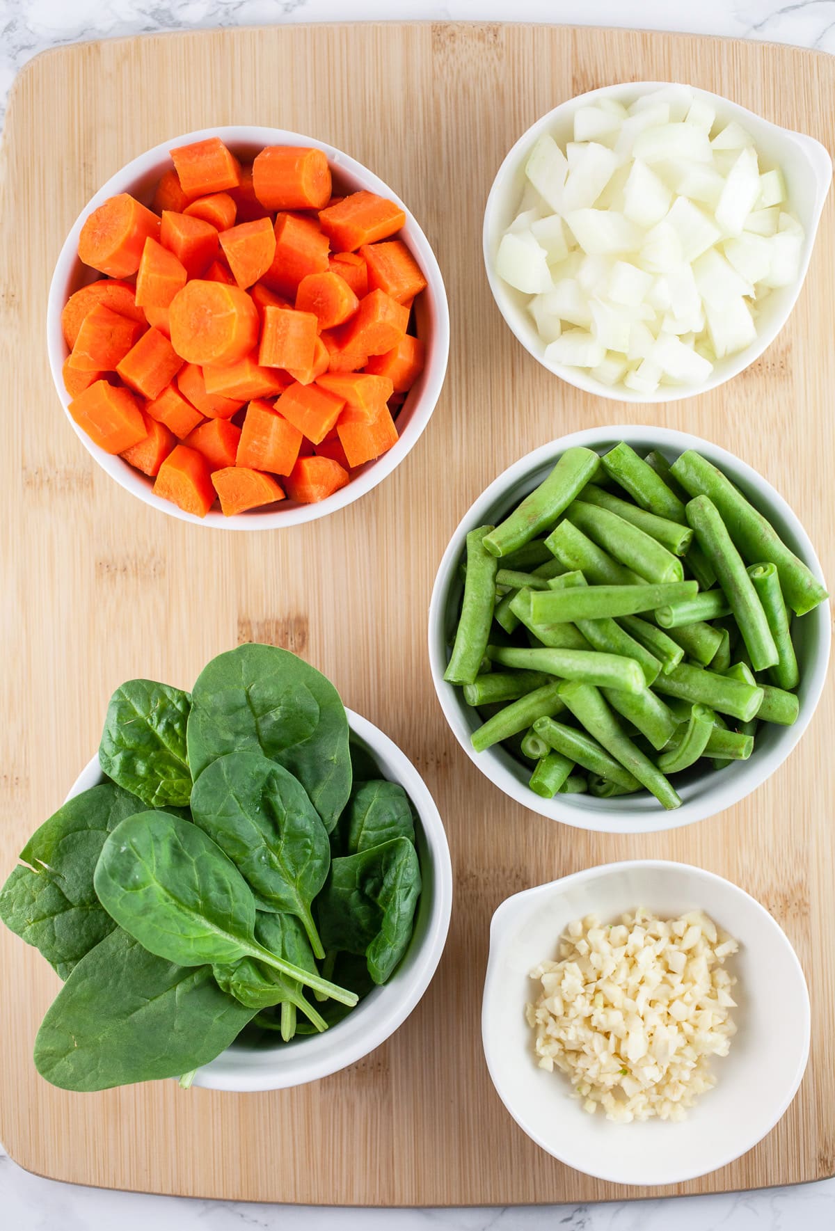 Minced garlic and onions, chopped carrots and green beans, and fresh spinach on wooden cutting board.