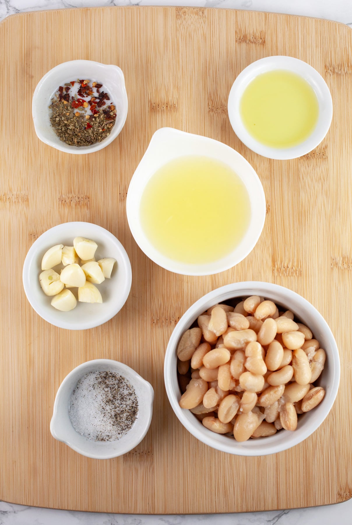 Cannellini beans, garlic, kosher salt, pepper, Italian seasoning, red pepper flakes, lemon juice, and olive oil in small white bowls on wooden cutting board.