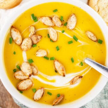 Butternut squash soup with pumpkin seeds and minced chives in white bowl with spoon.