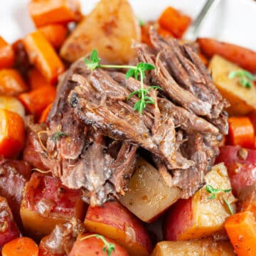 Pot roast on top of braised potatoes and carrots in white bowl with fork.